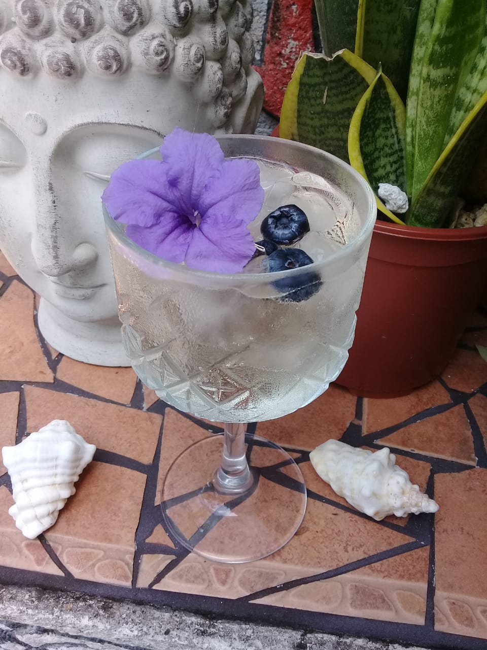 Refreshing cocktail with ice, blueberries, and a purple flower garnish beside Buddha statue.